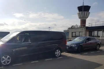 Van noir et berline grise devant une tour de contrôle d’aéroport.